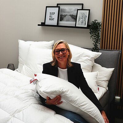 The new managing director sits relaxed on a bed, holding a pillow and smiling warmly at the camera.