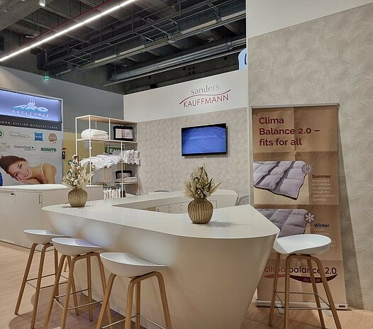 Messestand auf der Heimtextil 2024 in Cremetönen. Regale mit ausgestellten Bettdecken und Kissen.