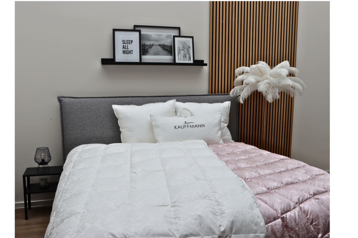 A modern bedroom with a grey bed, a white and pink duvet, a decorative feather lamp and a wall shelf with pictures above the bed.