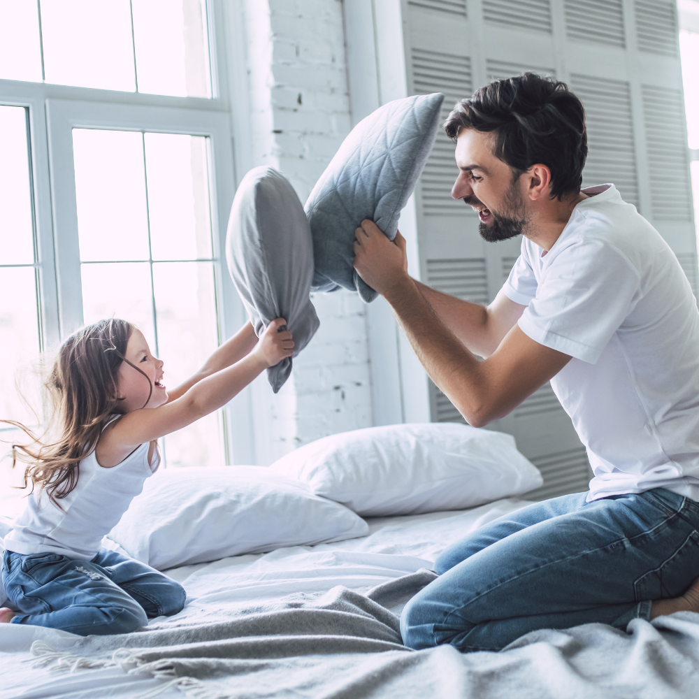 Vater und Tochter sitzen im Bett und spielen fröhlich mit Kissen während einer Kissenschlacht.
